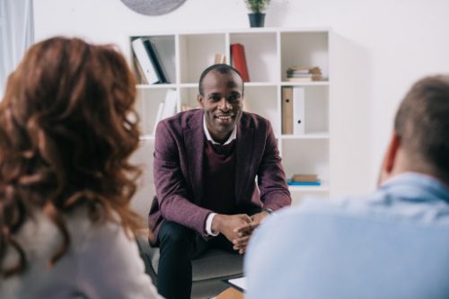 A couple on a therapy session with their phycologist sitting on a chair in front of them smiling with his finger crossed divorce mediation