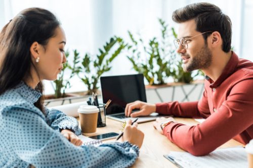 man smiling while looking at the woman with a laptop on his side, woman looking at the table seriously with a pen on her hands and a coffee on her side postnuptial agreements